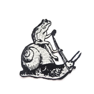 May include: Black and white embroidered patch featuring a frog riding a snail. The frog is holding a stick, and the snail has its antennae extended. The patch is on a white background.