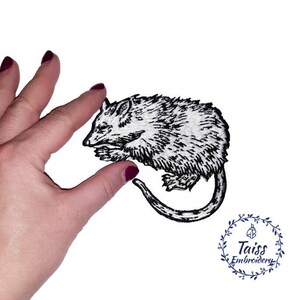 May include: Black and white embroidered iron-on patch of a curled up possum. The patch is on a white background and is being held by a hand.