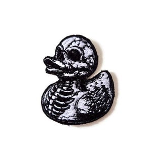 May include: A black and white embroidered patch featuring a rubber duck design, with a skull and ribcage motif. The patch has a detailed, stitched appearance, with a white design on a black background. The duck is facing left, with its beak open.