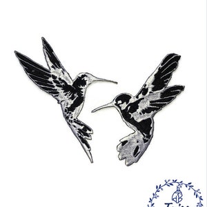 Puede incluir: Dos parches bordados de colibrí en blanco y negro. Los colibríes se miran, con las alas extendidas. Los parches están sobre un fondo blanco.