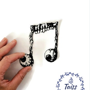 May include: Black and white embroidered iron-on patch of a musical note with two eyeballs. The note is shaped like a traditional musical note with a jagged edge at the top. The eyeballs are detailed with pupils and eyelids.