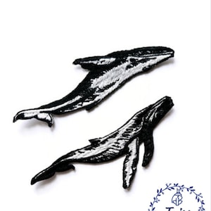 Könnte beinhalten: Zwei gestickte Wal-Patches in Schwarzweiß. Die Wale sind in entgegengesetzte Richtungen gerichtet und befinden sich in Schwimmposition.