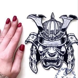 Peut inclure: Un &eacute;cusson brod&eacute; en noir et blanc repr&eacute;sentant un casque de samoura&iuml; avec une expression f&eacute;roce. L'&eacute;cusson est sur un fond blanc et est entour&eacute; d'une main.