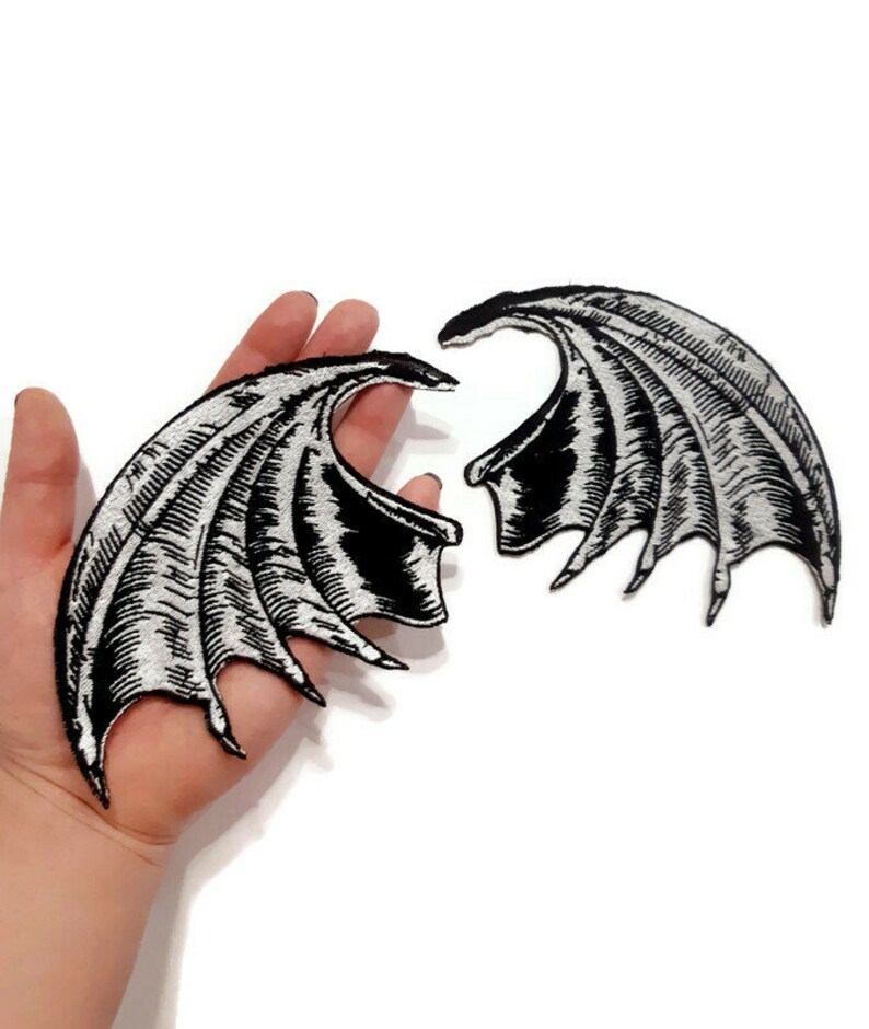 K&ouml;nnte beinhalten: Zwei schwarze und silberne gestickte B&uuml;gelbilder in Fl&uuml;gelform. Die Fl&uuml;gel sind mit einem strukturierten, schuppigen Design verziert.