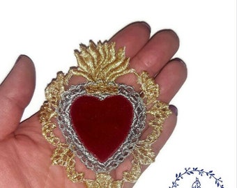 Puede incluir: Corazón bordado en oro y plata con un centro de terciopelo rojo. El corazón está rodeado de un diseño floral decorativo.