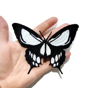 May include: Black and white embroidered iron-on patch of a butterfly with a skull face. The butterfly has white eyes and a white skull with teeth.