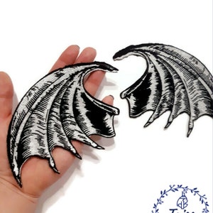 K&ouml;nnte beinhalten: Zwei schwarze und silberne gestickte B&uuml;gelbilder in Fl&uuml;gelform. Die Fl&uuml;gel sind mit einem strukturierten Design verziert und haben eine spitze Form.