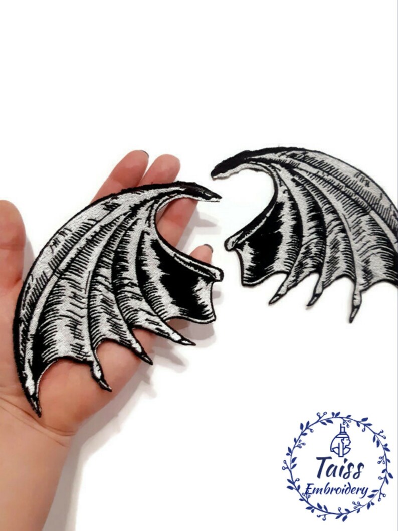 K&ouml;nnte beinhalten: Zwei schwarze und silberne gestickte B&uuml;gelbilder in Fl&uuml;gelform. Die Fl&uuml;gel sind mit einem strukturierten Design verziert und haben eine spitze Form.