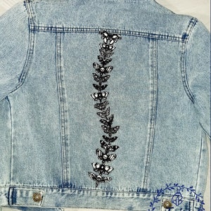 Peut inclure: Une veste en jean bleu clair avec un motif brodé noir et blanc de papillons et de crânes sur le dos. La broderie est en ligne verticale et est centrée sur le dos de la veste.