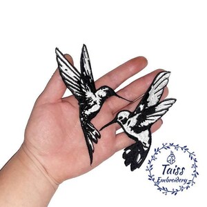 K&ouml;nnte beinhalten: Zwei gestickte Kolibri-Patches in Schwarz und Wei&szlig;. Die Patches befinden sich auf einer Hand, mit dem Text "Taiss Embroidery" in einem Kreis darunter.