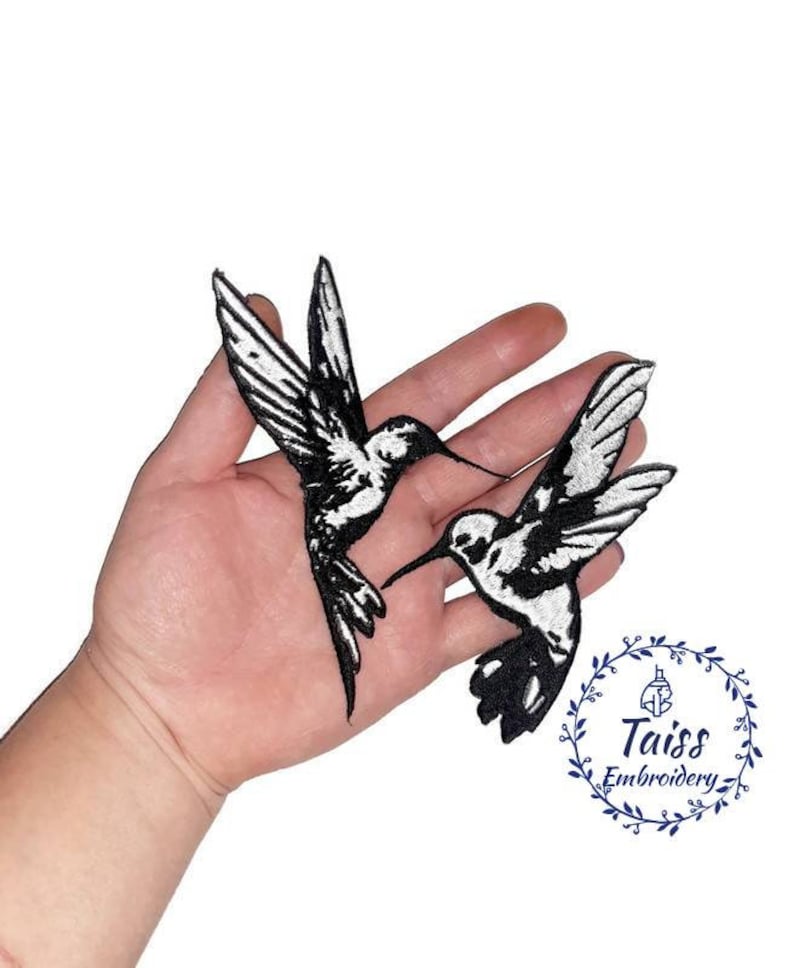 K&ouml;nnte beinhalten: Zwei gestickte Kolibri-Patches in Schwarz und Wei&szlig;. Die Patches befinden sich auf einer Hand, mit dem Text "Taiss Embroidery" in einem Kreis darunter.