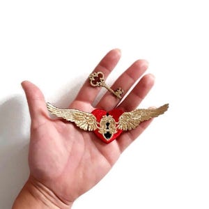 May include: A red heart-shaped patch with gold embroidered wings and a keyhole design. A small gold-colored key is also present. The patch and key are likely for decorative purposes.