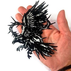 May include: Black and white embroidered patch depicting a raven in flight. The patch has a detailed design with a textured appearance, suitable for application on clothing or accessories.