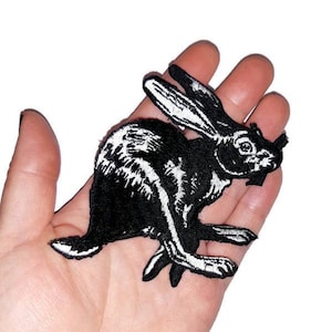 May include: Black and white embroidered iron-on patch of a rabbit running. The rabbit is outlined in white and filled in with black.