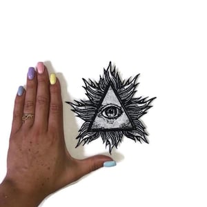 Peut inclure: Écusson noir et blanc représentant un œil dans un triangle, entouré d'un motif de flammes stylisées. Ce patch, destiné aux vêtements ou accessoires, a une thématique mystique ou occulte.