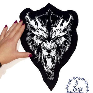 May include: Black embroidered patch featuring a white wolf's head design. The wolf has a fierce expression and a long, pointed snout. The patch is shaped like a triangle and has a raw edge.