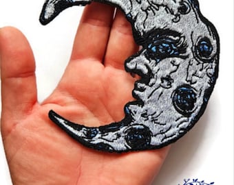 Puede incluir: Un parche bordado en forma de media luna con un diseño detallado que presenta una cara y cráteres. La luna es blanca con acentos azules y costuras negras.