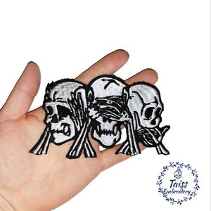 Könnte beinhalten: Drei gestickte Bügelbilder mit einem Trio von Schädeln, jeder mit einer Hand, die einen ihrer Sinne bedeckt: sehen, hören und sprechen. Die Schädel sind weiß mit schwarzen Umrissen und die Hände sind schwarz.