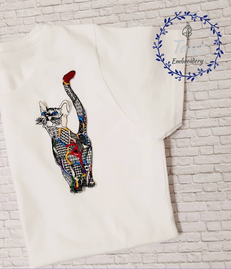May include: White t-shirt with a colorful embroidered cat design. The cat is facing away from the viewer and has a long tail. The embroidery is detailed and intricate, with a variety of colors and patterns.