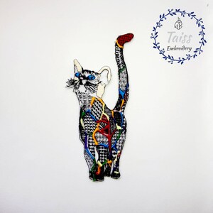 May include: A colorful embroidered patch of a cat with a black and white patterned body and a red tail. The cat has blue eyes and a white face. The patch is on a white background.