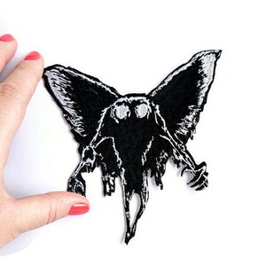 May include: Black and white embroidered patch of a creature with large eyes and wings. The creature has a menacing expression and appears to be flying.