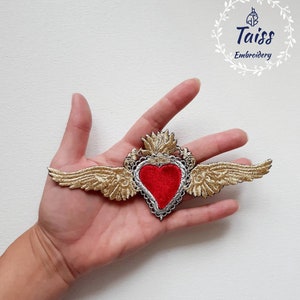 May include: Gold embroidered patch with a red velvet heart and wings. The patch is held in a hand. The text "Taiss Embroidery" is in the upper right corner of the image.