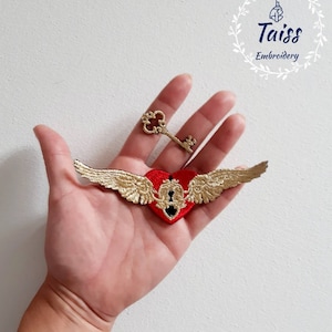 Puede incluir: Un corazón rojo con alas doradas y un ojo de cerradura, con una llave dorada separada, bordado sobre un fondo blanco.