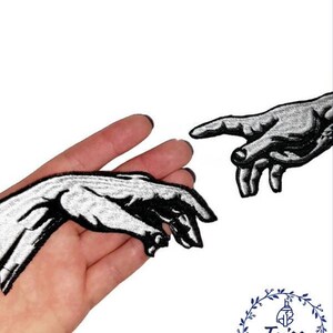 Pu&ograve; includere: Due toppe ricamate in bianco e nero di mani che si raggiungono a vicenda. Le toppe sono su uno sfondo bianco.