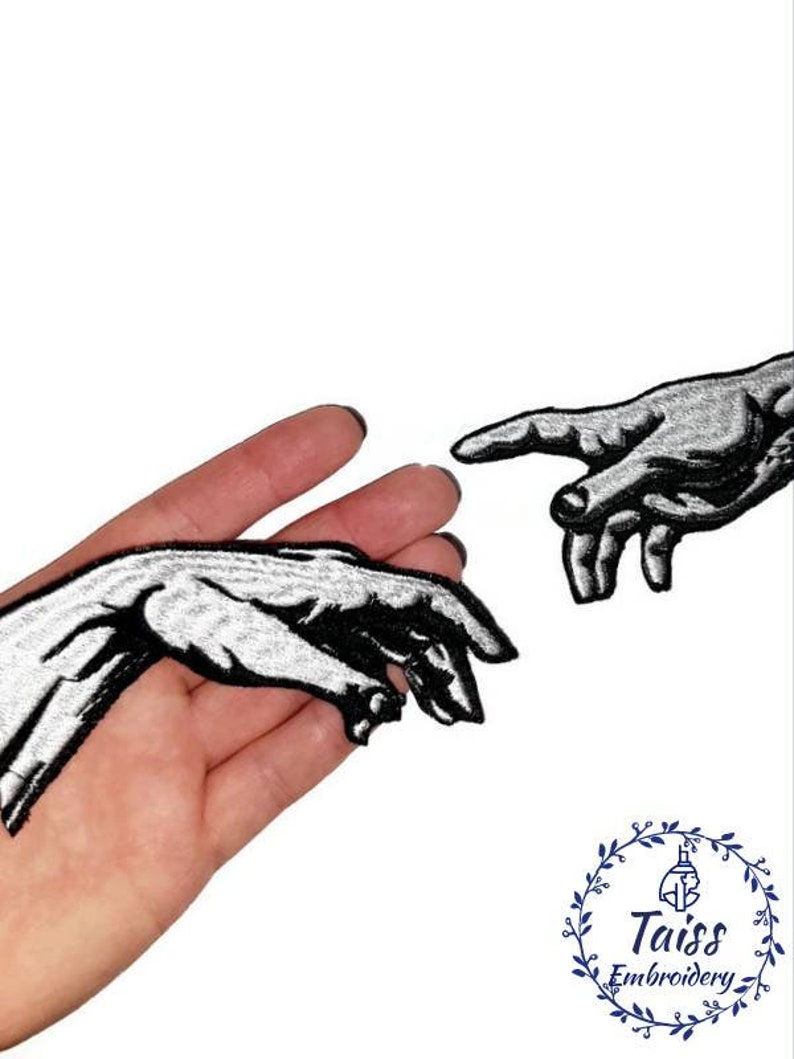 Pu&ograve; includere: Due toppe ricamate in bianco e nero di mani che si raggiungono a vicenda. Le toppe sono su uno sfondo bianco.