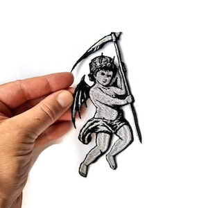 May include: Embroidered patch featuring a cherubic figure with wings, a crown, and a scythe. The design is in shades of gray and black, with detailed stitching. The patch is held against a white background.