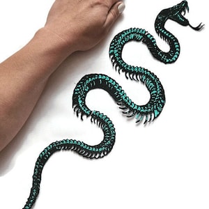 May include: A black and teal snake-shaped patch with intricate detailing. The patch features a detailed design with a snake's head and a long, winding body. The snake is teal with black accents. The patch is on a white background.