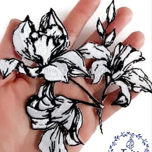 May include: White embroidered flower patch with black outlines. The patch is on a hand and features a delicate floral design.