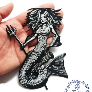 Może przedstawiać: Czarno-biała, haftowana naszywka z syreną trzymającą trójząb. Syrena ma długie, powiewające włosy i szczegółowy rybi ogon. Naszywka jest trzymana w dłoni na białym tle. Naszywka pochodzi z Taiss Embroidery.