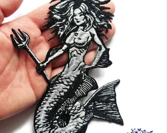 Pode incluir: Patch bordado a preto e branco de uma sereia segurando um tridente. A sereia tem cabelo comprido e esvoaçante e uma cauda de peixe detalhada. O patch é segurado em uma mão contra um fundo branco. O patch é da Taiss Embroidery.