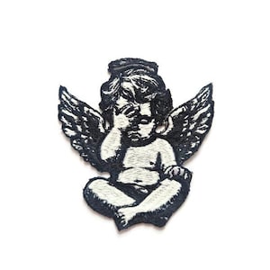 May include: Embroidered patch featuring a cherub with wings, in a seated pose, covering its eyes. The design is black and white, with detailed stitching. The patch is likely for clothing or accessories.