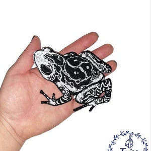 Może przedstawiać: Czarno-biała haftowana naszywka z motywem żaby. Żaba ma szczegółowy wzór z misternymi wzorami.