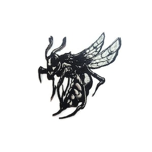 May include: A black and white insect-like patch with detailed wings, legs, and antennae. The patch has a textured appearance and is set against a plain white background. The design is intricate and stylized.