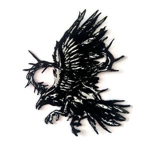 May include: A black and white embroidered patch featuring a raven in flight, encircled by a thorny wreath. The raven's wings are spread, and its talons are extended. The patch is likely designed to be sewn onto clothing or accessories.