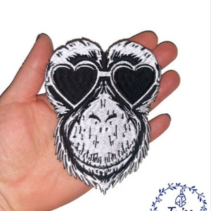May include: Black and white embroidered patch of a monkey face wearing heart-shaped sunglasses. The patch has detailed stitching and a playful design. Ideal for customising clothing or accessories. The patch is a fun, quirky item.