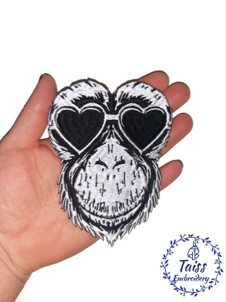 K&ouml;nnte beinhalten: Schwarz-wei&szlig;er, gestickter Aufn&auml;her mit einem Affengesicht mit herzf&ouml;rmiger Sonnenbrille. Der Aufn&auml;her hat detaillierte Stickereien und ein verspieltes Design. Ideal zum Anpassen von Kleidung oder Accessoires. Ein lustiges, skurriles Produkt.