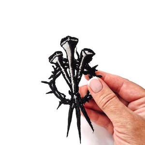 May include: Black embroidered patch featuring three nails and a thorny wreath. The design is detailed with white highlights, creating a striking contrast against the black. This patch is suitable for jackets, bags, or other fabric items.