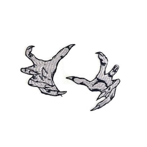 May include: Two embroidered patches of clawed hands with sharp nails, in a silver color, on a white background.
