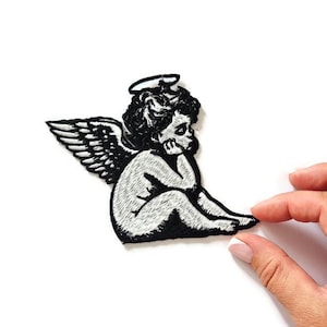 Puede incluir: Parche bordado en blanco y negro con un diseño de querubín. El querubín está representado con alas y un halo, sentado en una pose pensativa. Adecuado para ropa o accesorios.