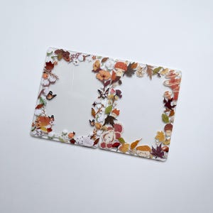 May include: A clear, rectangular card holder with a decorative border of autumn-themed illustrations. The border features colorful leaves, mushrooms, flowers, and butterflies. The card holder is open, with two sections.