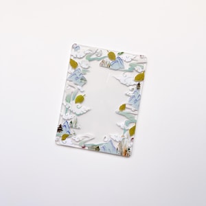 Pode incluir: Um porta-retratos de acrílico transparente com uma borda decorativa que representa uma paisagem azul e verde com folhas amarelas. O porta-retratos é perfeito para exibir uma foto ou obra de arte favorita.