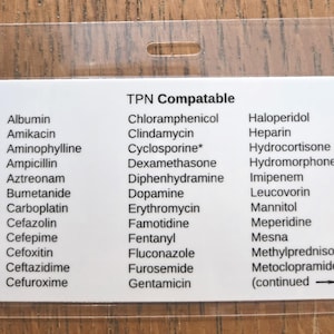 Puede incluir: Una tarjeta de plástico blanco con una lista de medicamentos, incluyendo Albumin, Amikacin y Cloramfenicol, bajo el título "TPN Compatable". La tarjeta tiene un agujero en la parte superior y está sobre un fondo de madera.