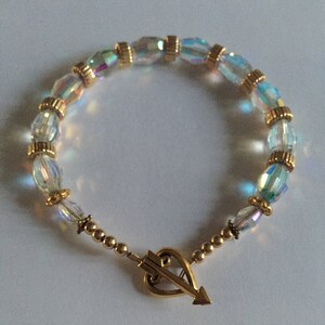 May include: A gold heart charm with an arrow through it hangs from a bracelet made of clear iridescent glass beads and gold beads.