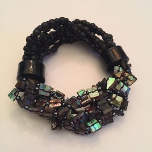May include: A black beaded bracelet with iridescent abalone shell accents. The bracelet is made with multiple strands of black beads and features a unique design with the abalone shell pieces clustered together.