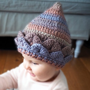 K&ouml;nnte beinhalten: Ein Baby tr&auml;gt eine geh&auml;kelte M&uuml;tze mit einer spitzen Krone und einem gewellten Rand in den Farben Lila, Braun und Rosa.