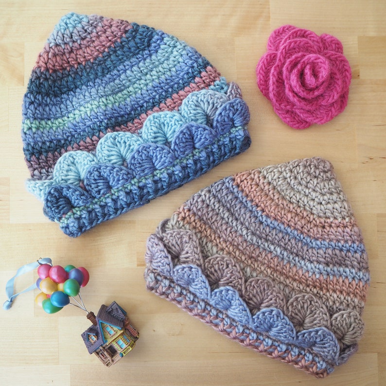 K&ouml;nnte beinhalten: Zwei geh&auml;kelte M&uuml;tzen mit einem gewellten Rand in Blaut&ouml;nen, Rosa und Braun. Die M&uuml;tzen sind mit einem strukturierten Stich gearbeitet und haben einen einzigartigen, handgefertigten Look.